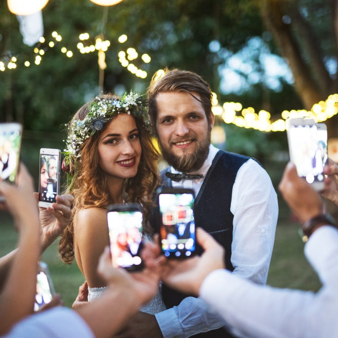 Guests with smartphones taking photo of bride and groom at wedding reception outside.