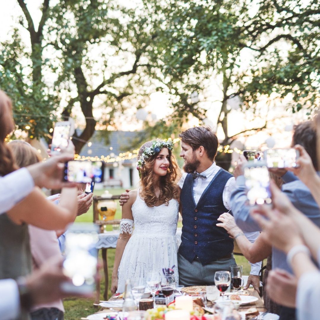 Guests with smartphones taking photo of bride and groom at wedding reception outside.